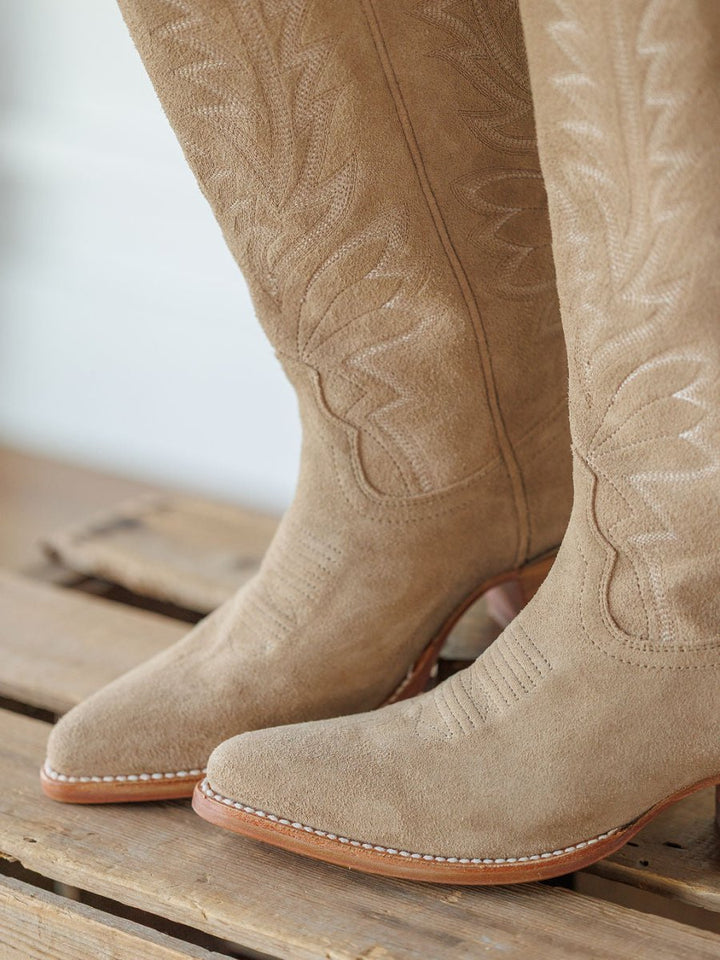 Close-up of Savannah Sevens western life{&}style's The Abby Boot {Driftwood}, featuring premium suede, decorative stitching, pointed toes, and wooden soles, standing on a wooden surface. Boots handmade by Tecovas.