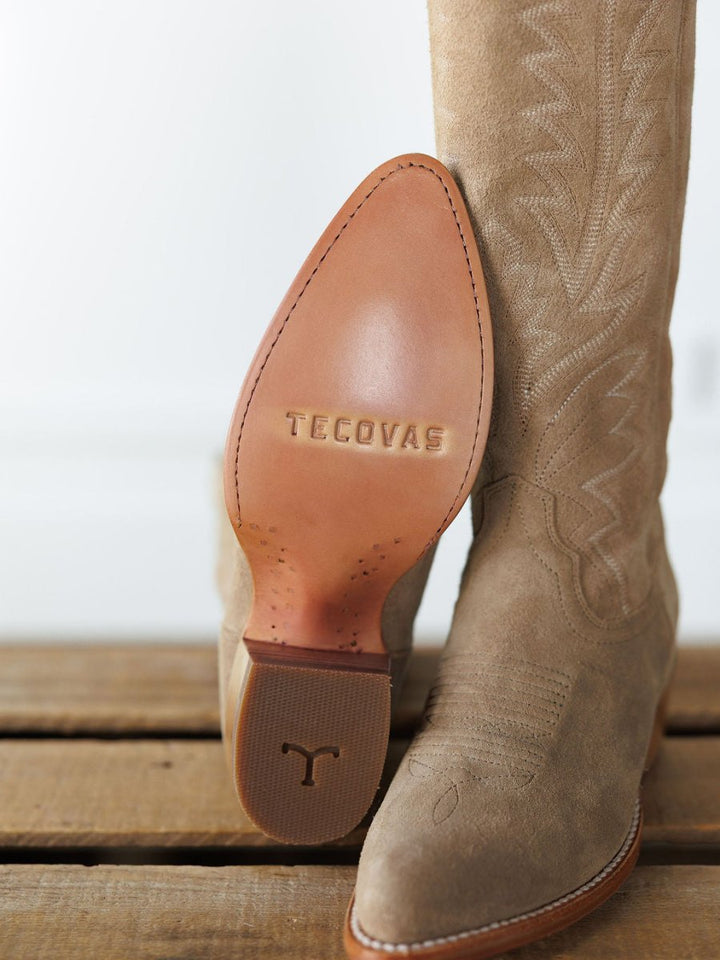 A close-up of Savannah Sevens western life{&}style's The Abby Boot {Driftwood}: one boot stands upright, the other sole-up. Premium suede leather, textured heel, and decorative stitching highlight its Western style. Boots handmade by Tecovas.
