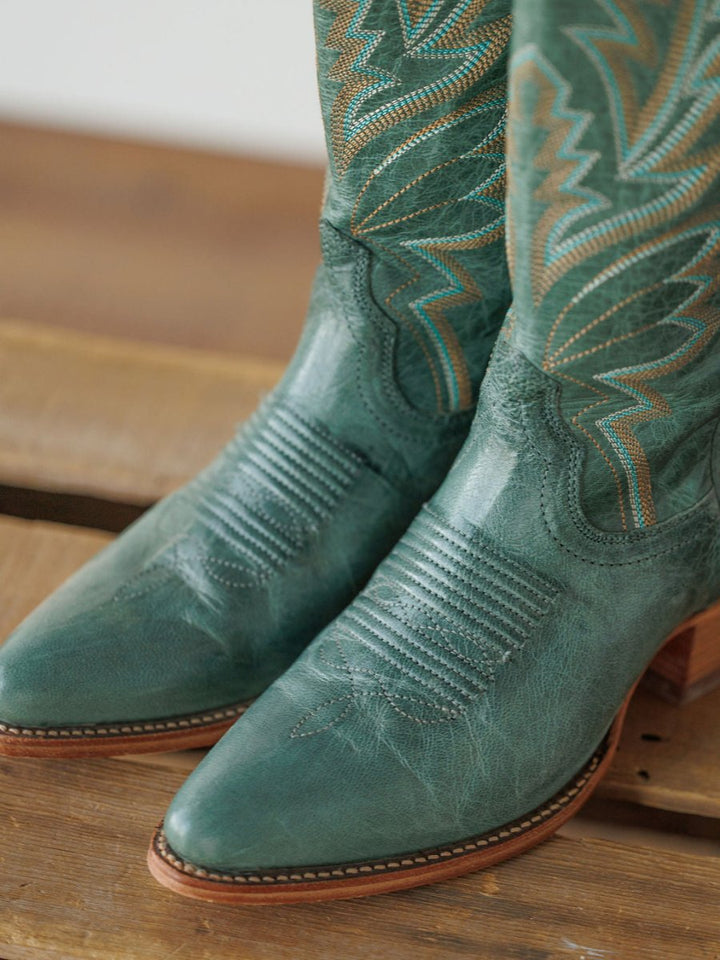 A close-up of The Abby Boot {Sagebrush} by Savannah Sevens western life{&}style, featuring decorative stitching and wooden heels, resting on a wooden surface. Boots handmade by Tecovas.