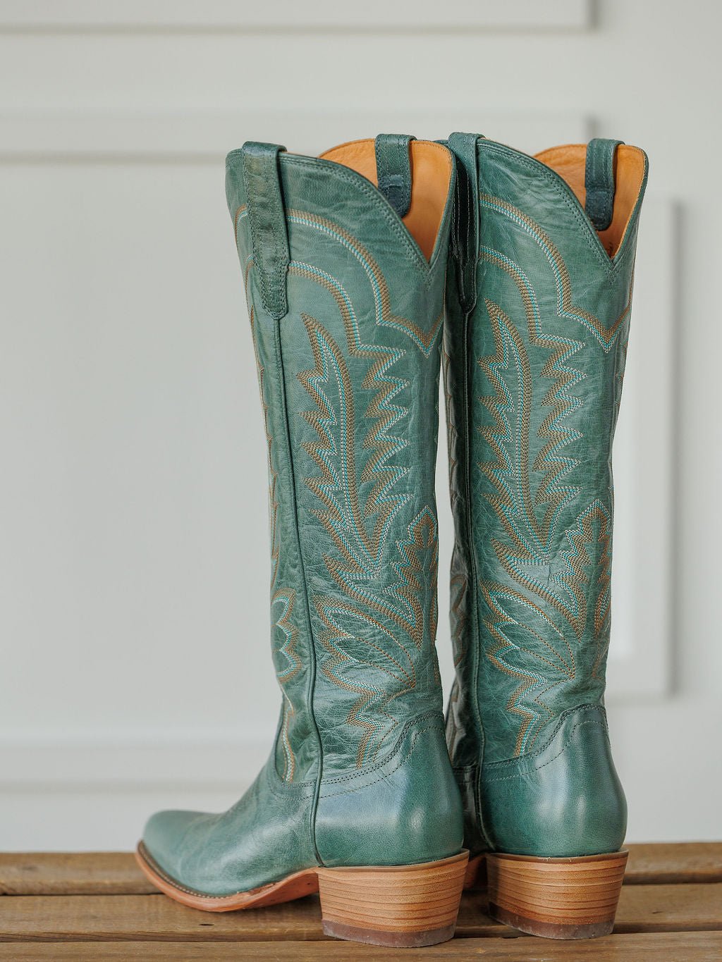 A pair of tall, sage green The Abby Boot {Sagebrush} by Savannah Sevens western life{&}style with decorative stitching and tan soles, standing on a wooden surface against a white background. Boots handmade by Tecovas.