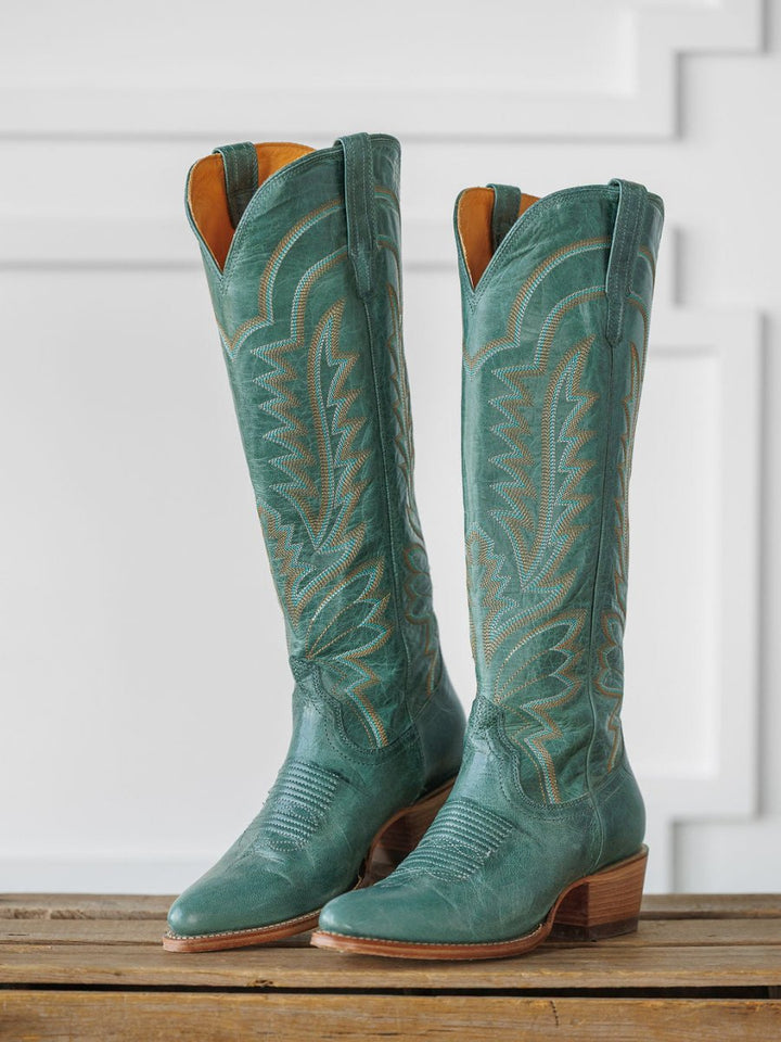 A pair of tall, sagebrush-green "The Abby Boot" by Savannah Sevens western life{&}style with decorative stitching and wooden heels, displayed upright on a wooden surface against a white paneled background. Boots handmade by Tecovas.