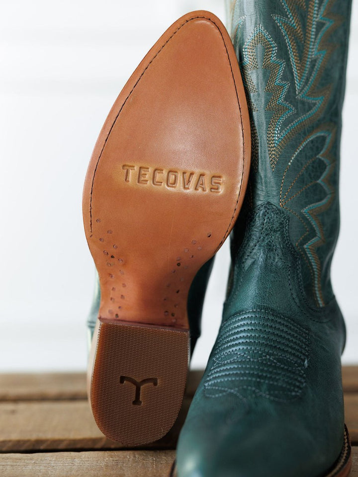 A close-up of Savannah Sevens western life{&}style’s The Abby Boot {Sagebrush}, one upright and one showing the tan leather sole. The boots are dark green with decorative stitching, displayed on a wooden surface. Boots handmade by Tecovas.