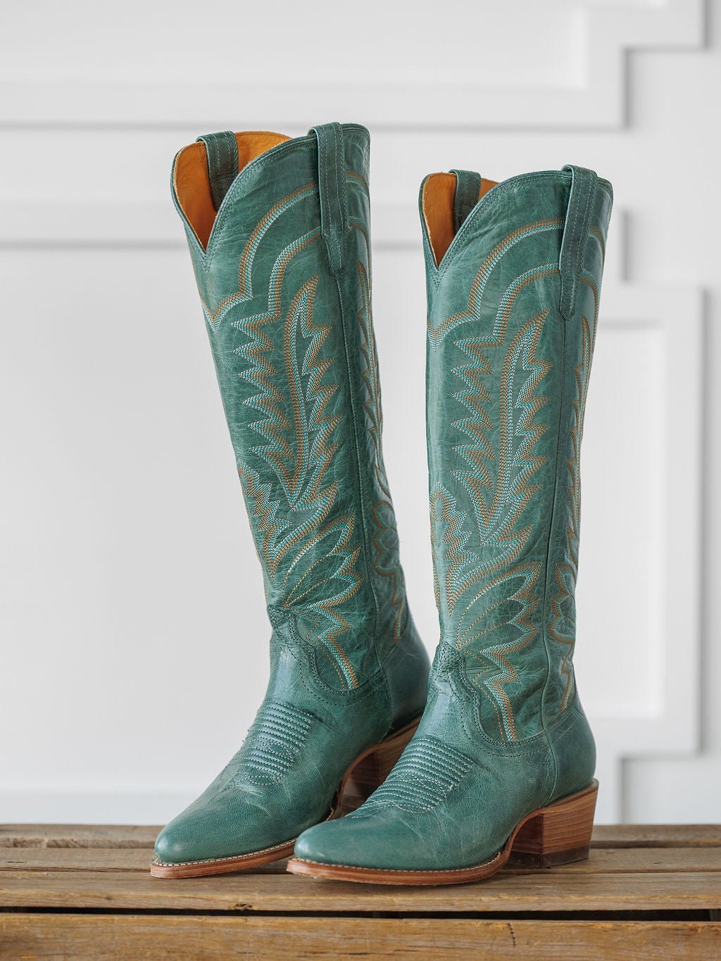 A pair of tall, sagebrush-green "The Abby Boot" by Savannah Sevens western life{&}style with decorative stitching and wooden heels, displayed upright on a wooden surface against a white paneled background. Boots handmade by Tecovas.