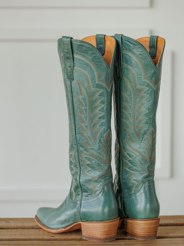 A pair of tall, sage green The Abby Boot {Sagebrush} by Savannah Sevens western life{&}style with decorative stitching and tan soles, standing on a wooden surface against a white background. Boots handmade by Tecovas.