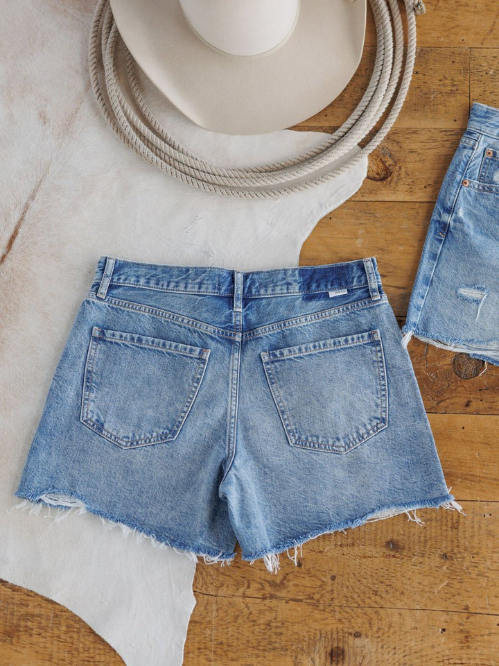 The Ace Shorts by Savannah Sevens western life{&}style—a pair of high rise, medium light wash denim shorts with a raw cut frayed hem—are displayed on a wooden floor beside a cowboy hat and cowhide rug. Another denim shorts pair is partially visible.