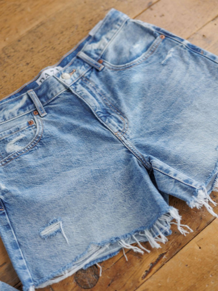 The Ace Shorts by Savannah Sevens western life{&}style are high rise, medium light wash denim shorts with a raw cut frayed hem and ripped details, shown lying flat on a wooden surface.