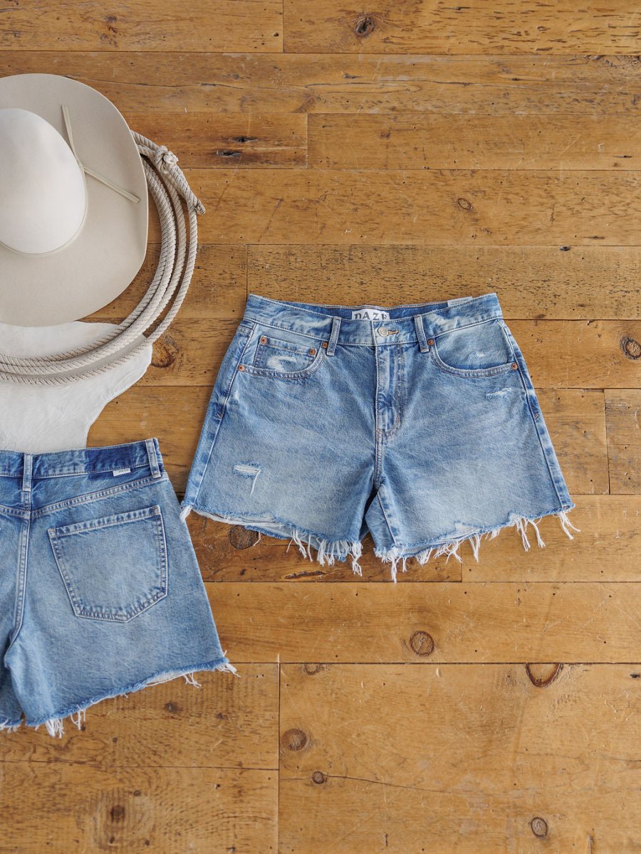 Two pairs of The Ace Shorts from Savannah Sevens western life{&}style, high rise denim with raw cut frayed hems in medium wash, are displayed on a wooden floor beside a white cowboy hat and coiled rope for a casual western vibe.