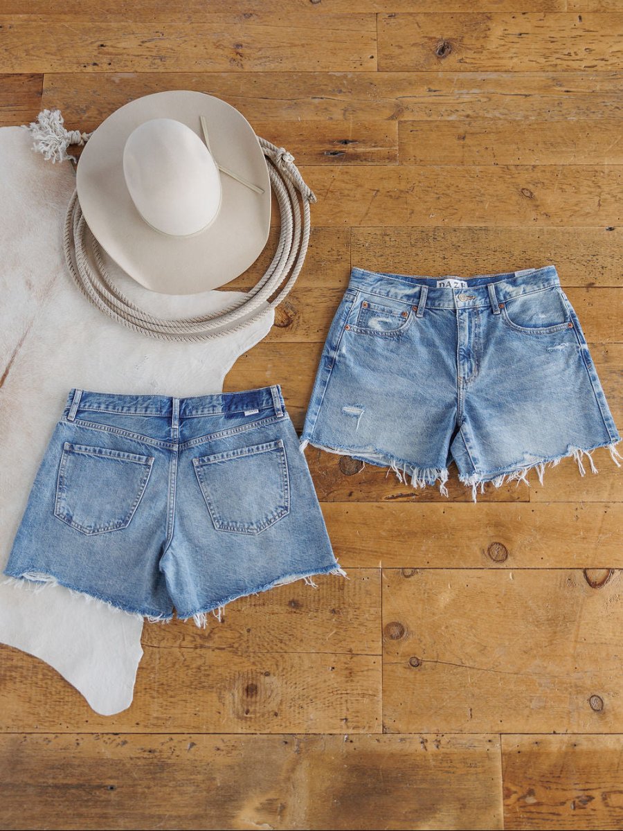 Flat lay of two pairs of The Ace Shorts by Savannah Sevens western life{&}style—high rise denim with a raw cut frayed hem in medium light wash—on wood floor beside a white wide-brim hat, coiled rope, and white cowhide rug.