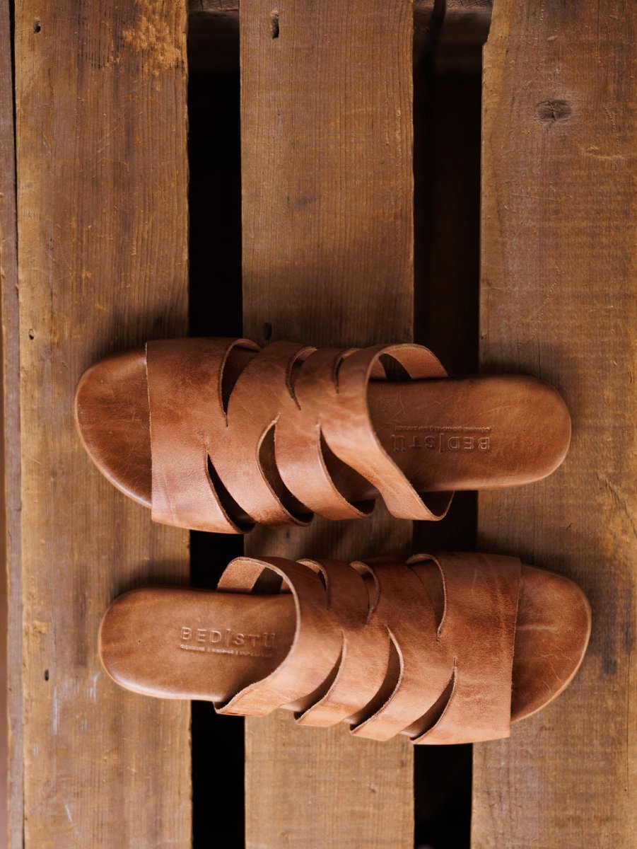 A pair of The Acomas tan leather strappy sandals from Savannah Sevens western life{&}style with crisscross straps are placed side by side on a rustic wooden surface. Sandals made by BedStu. 