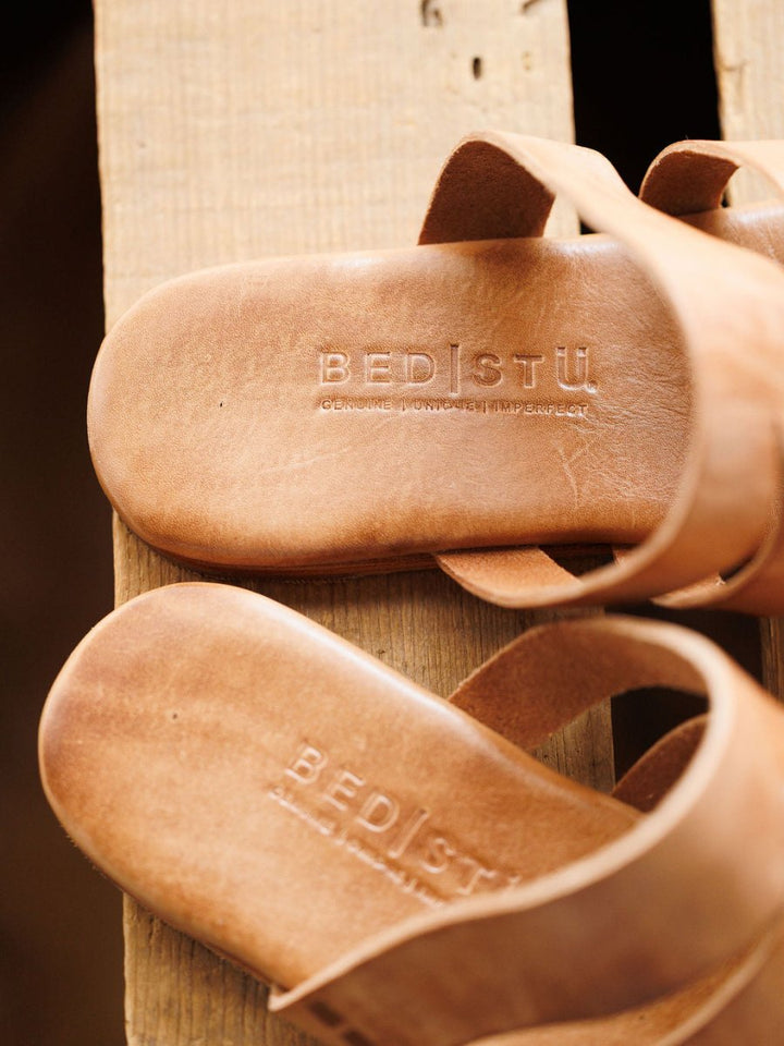 Close-up of two tan leather The Acomas sandals by Savannah Sevens western life{&}style on a wooden surface, with the Genuine Handcrafted Leather logo stamped on the insoles.