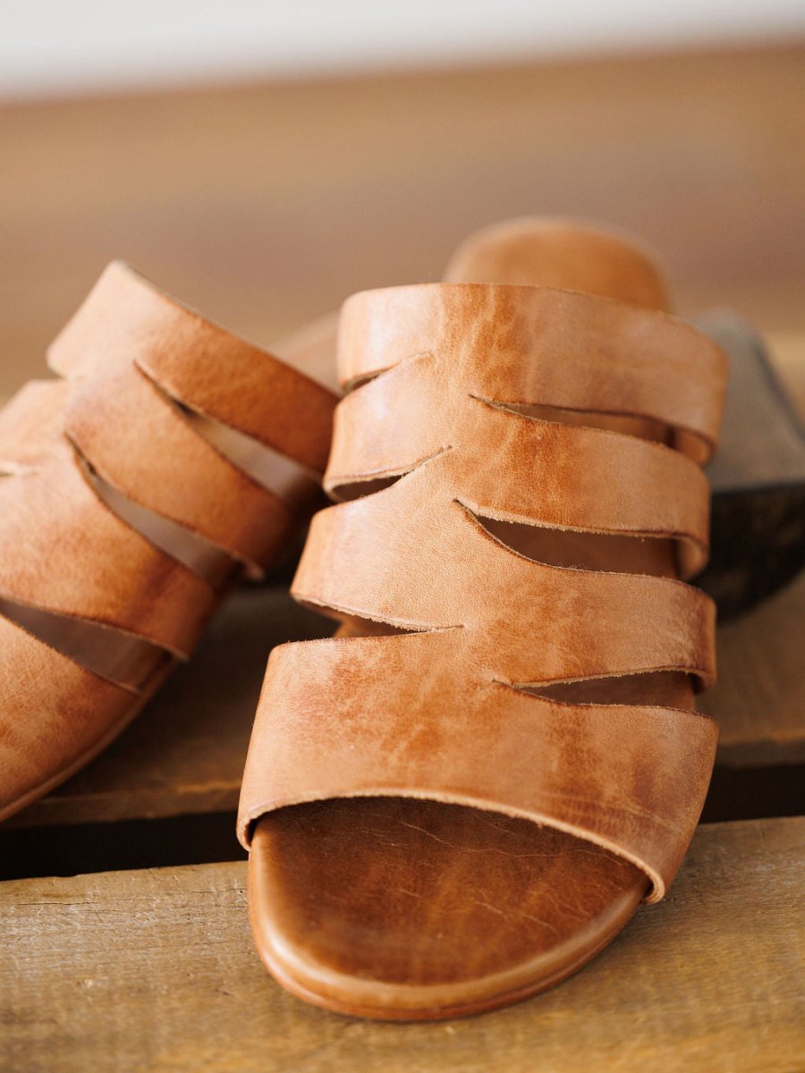 A close-up of The Acomas tan leather sandal by Savannah Sevens western life{&}style, featuring thick, overlapping straps and resting on a wooden surface.
