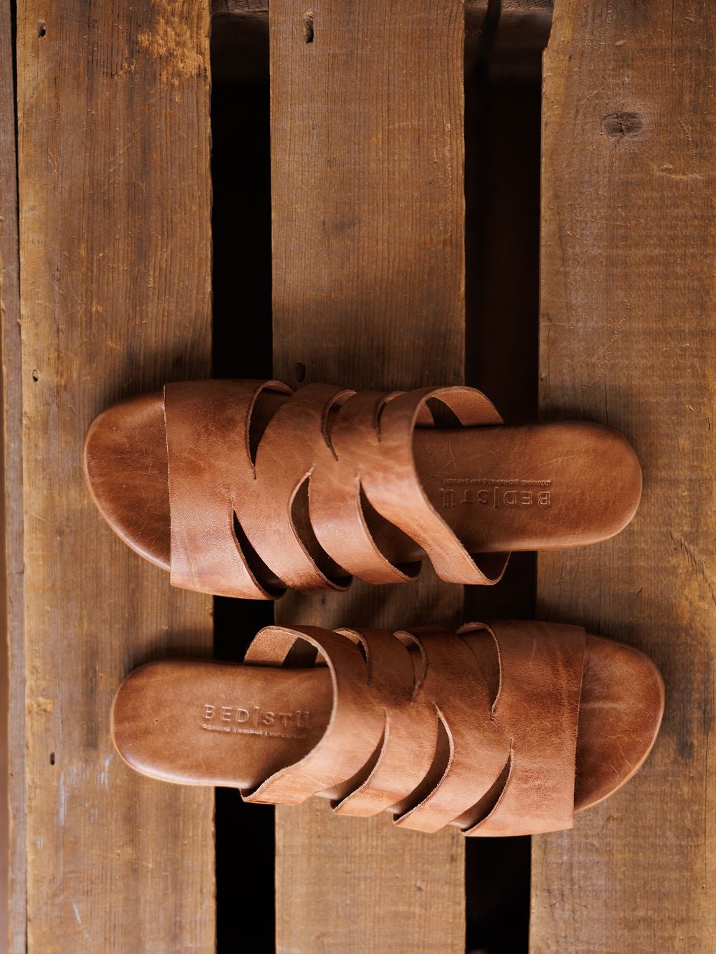 A pair of The Acomas tan leather strappy sandals from Savannah Sevens western life{&}style with crisscross straps are placed side by side on a rustic wooden surface. Sandals made by BedStu. 