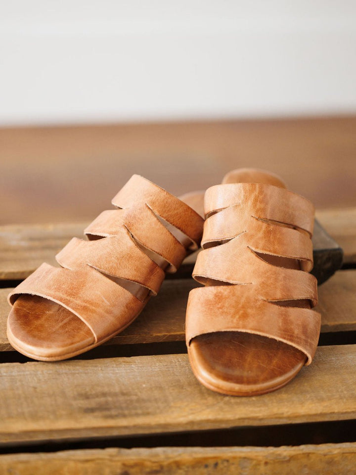 A pair of The Acomas tan leather sandals by Savannah Sevens western life{&}style with wide, crisscrossed straps displayed on a rustic wooden surface. Sandals made by BedStu. 