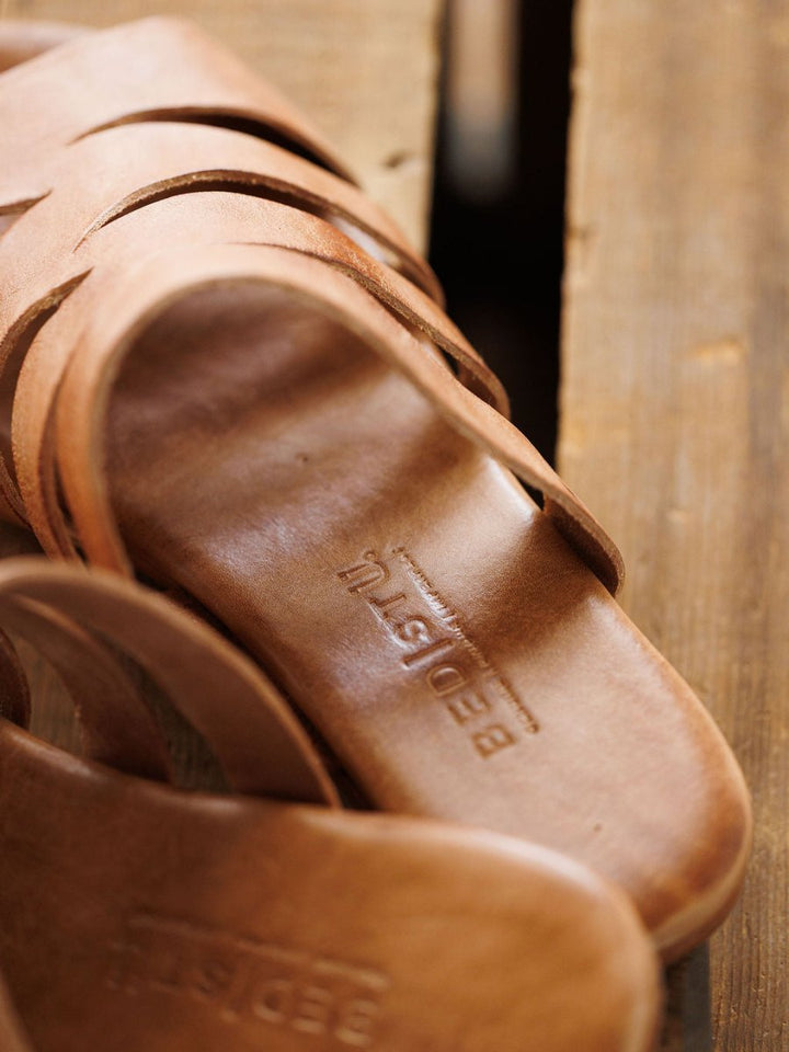 Close-up of The Acomas tan leather strappy sandal by Savannah Sevens western life{&}style on a wooden surface, with the brand name embossed inside the footbed.
