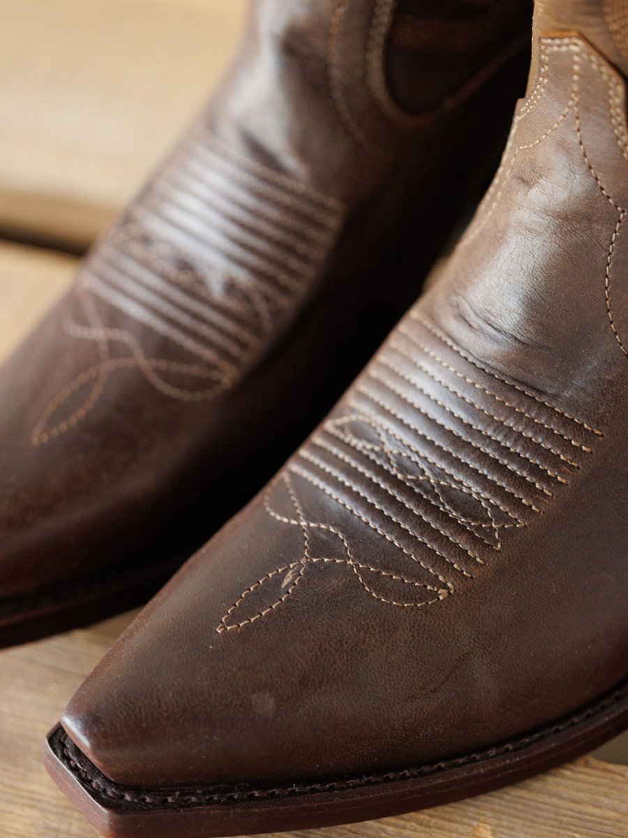 Close-up of the pointed snip toes on The Annie Boot {Cafe} by Savannah Sevens western life{&}style, crafted from brown premium goat leather with light decorative stitching, shown resting on a wooden surface. Boots handmade by Tecovas.