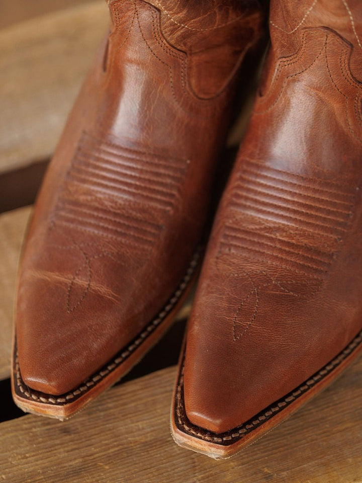 Close-up of the pointed toes of The Annie Boot {Scotch} by Savannah Sevens western life{&}style, featuring brown leather with decorative stitching and textured detailing, resting on a wooden surface. Boots handmade by Tecovas.