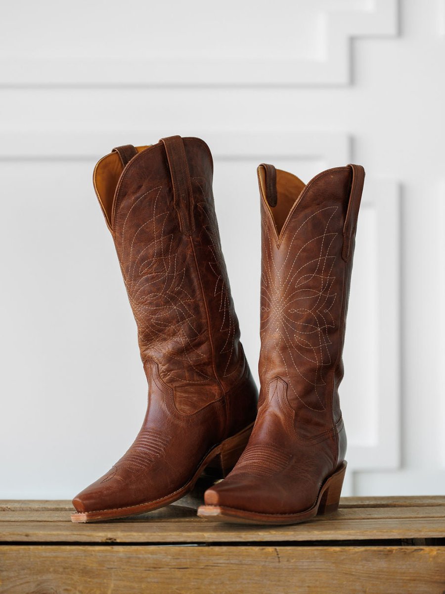 The Annie Boot {Scotch} by Savannah Sevens western life{&}style—a tall, brown Western leather boot with decorative stitching—is displayed side by side on a wooden surface against a white, textured background. Boots handmade by Tecovas.