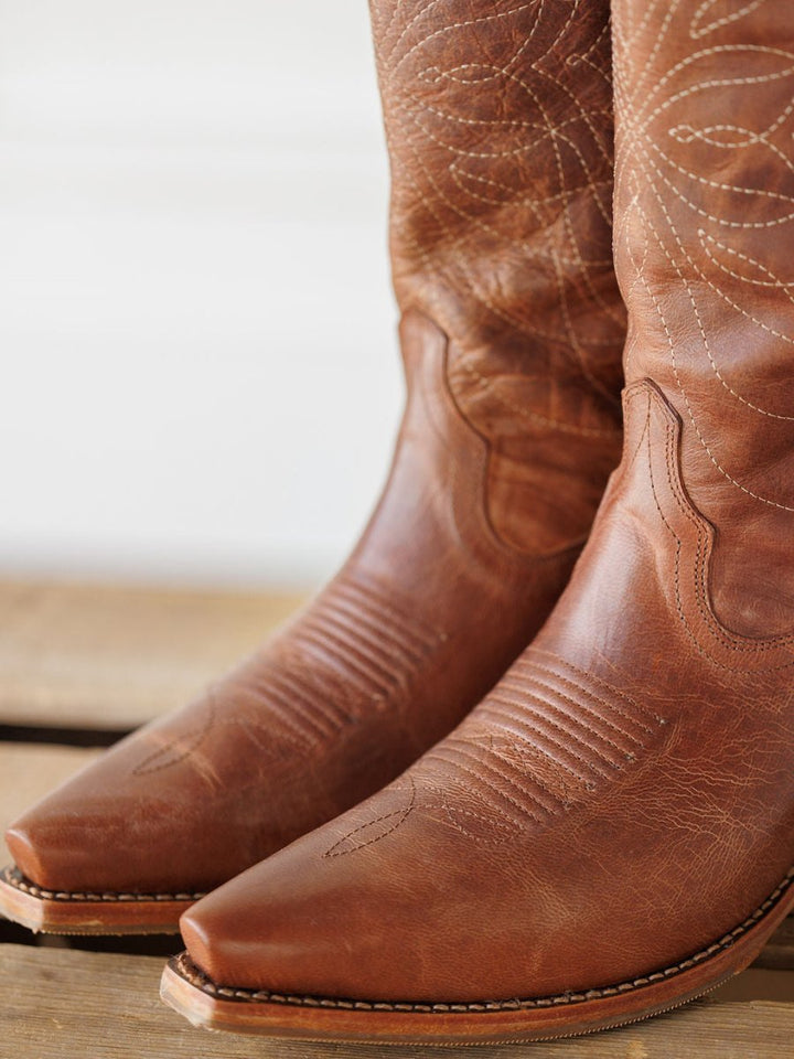 A close-up of Savannah Sevens western life{&}style’s The Annie Boot {Scotch}, featuring decorative stitching and a snip toe, shown resting on a wooden surface. Boots handmade by Tecovas.
