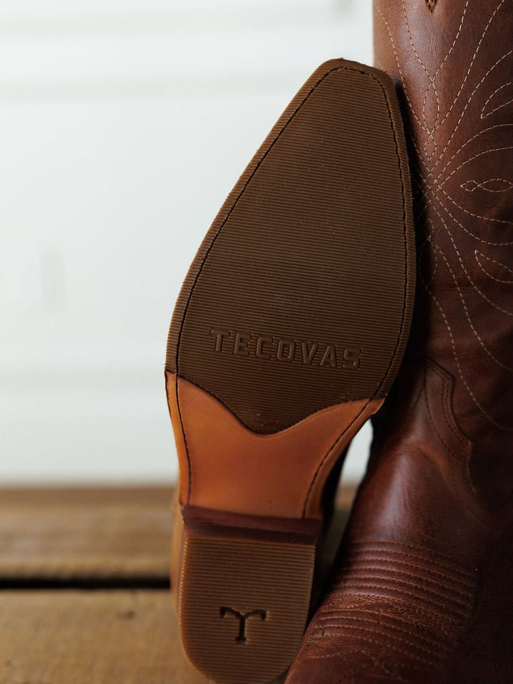 Close-up of the sole of The Annie Boot {Scotch} by Savannah Sevens western life{&}style, featuring the brand name embossed on the sole and decorative stitching on the leather upper. The snip toe boot rests on a wooden floor. Boots handmade by Tecovas.
