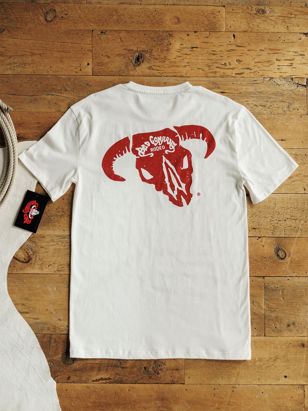 The Bad Company Men's Graphic Tee by Savannah Sevens western life{&}style—a white men's shirt with a bold red steer skull and "Rodeo Cowboys 2000" on the back—is shown flat on wood, with a black tag beside it.