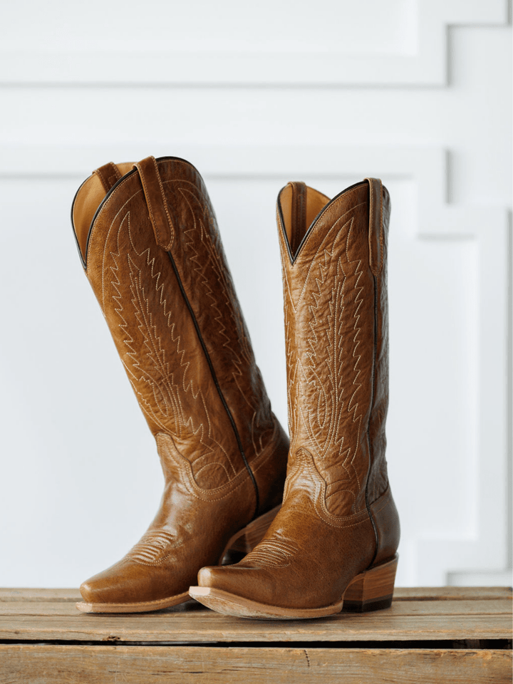 The Balcones boots from Savannah Sevens western life{&}style, tall and brown with intricate stitching, stand side by side on a wooden surface against a white decorative background. Made by Black Star. 