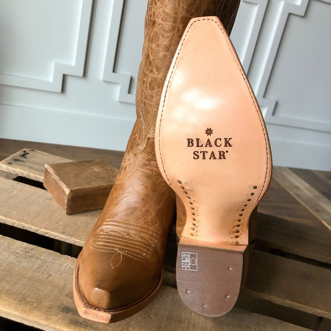A close-up of The Balcones pecan leather snip toe boot by Savannah Sevens western life{&}style on wood, showing the sole stamped with Black Star in black letters and white stitching details on the toe. Made by Black Star. 