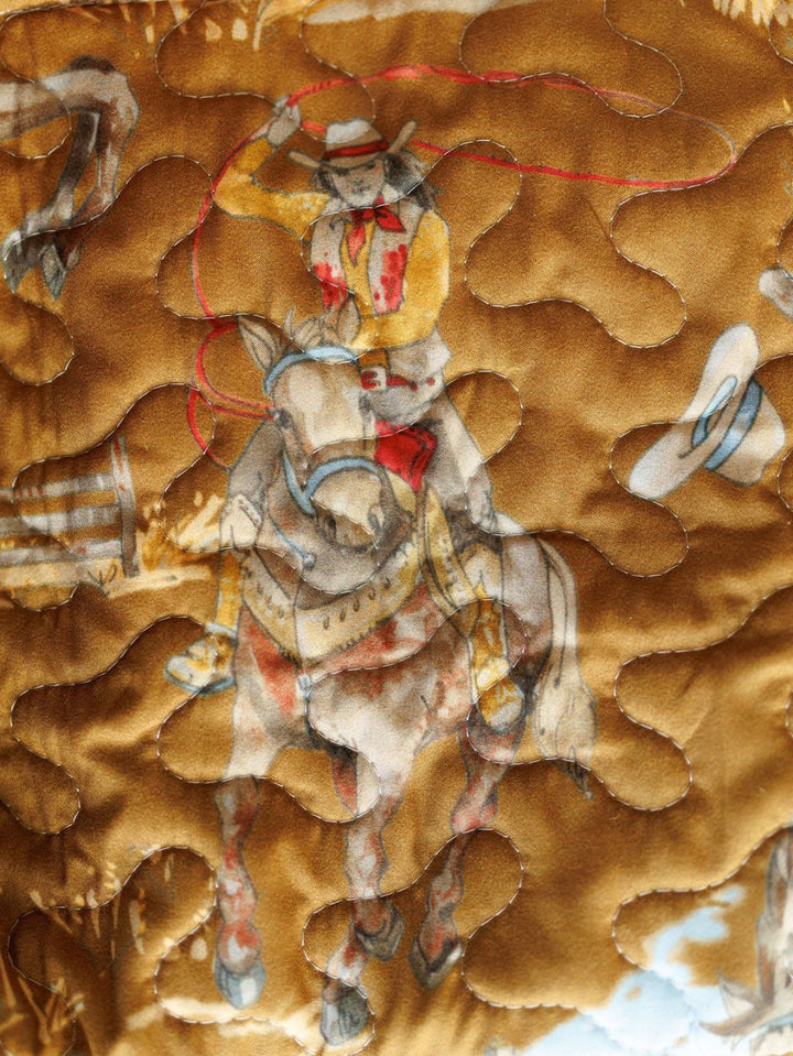 A close-up of The Big Sky Collection quilt by Savannah Sevens western life&style, featuring a painted cowboy on horseback, with a cowboy hat and barrel in the background and curved quilting stitches across the fabric.