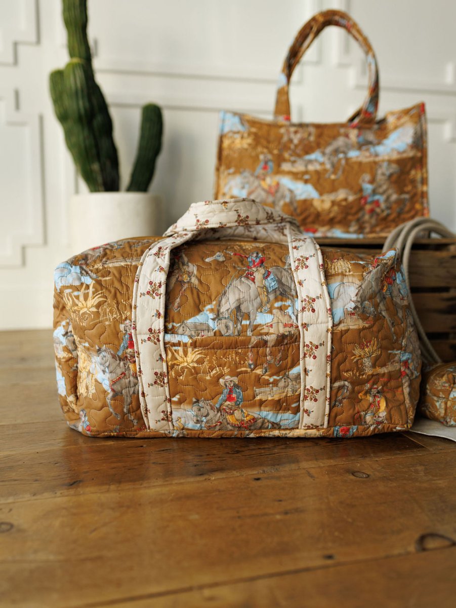 A quilted bag set from The Big Sky Collection by Savannah Sevens western life&style features a brown and blue scenic pattern, shown on a wooden floor with the matching tote and a potted cactus in the background.