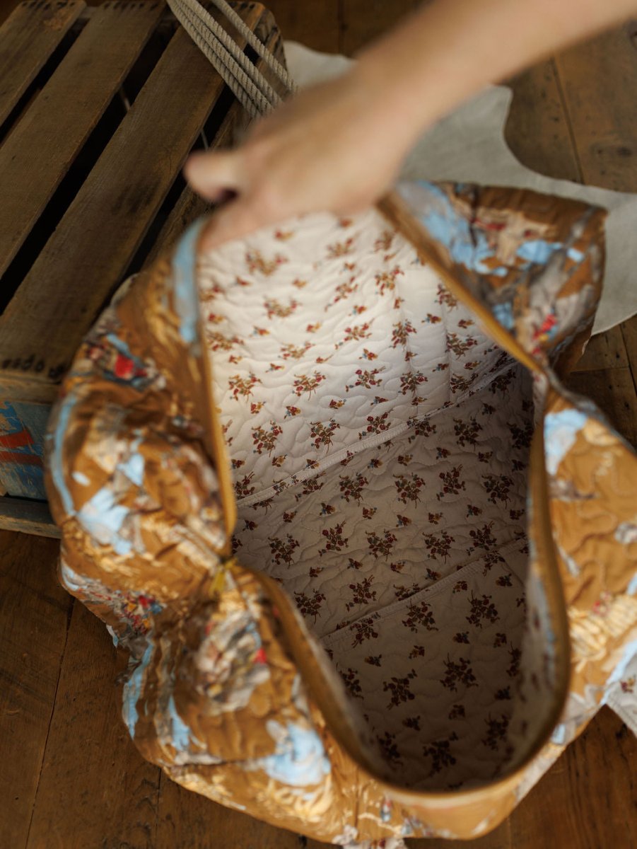 A hand displays The Big Sky Collection quilted bag set by Savannah Sevens western life & style, featuring a brown exterior with blue pattern, cream lining with red florals, resting on a wooden floor near a crate.