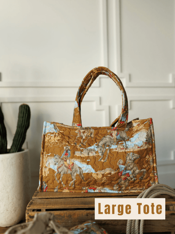 A quilted tote from The Big Sky Collection by Savannah Sevens western life&style, with a cowboy and horse print, rests on a crate by a potted cactus. "Large Tote" is boldly displayed at the bottom right.