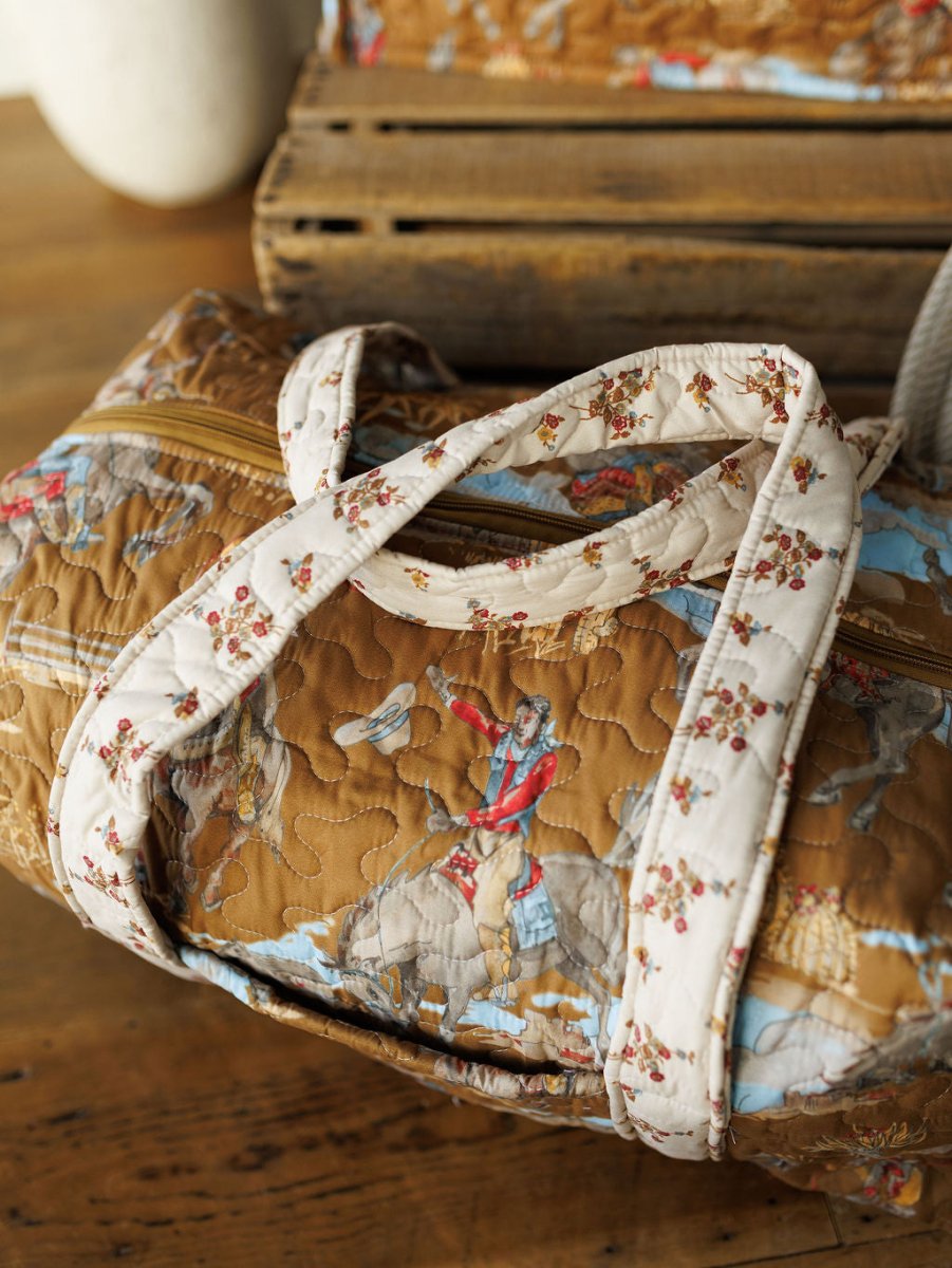 A quilted brown duffel bag from Savannah Sevens western life&style’s The Big Sky Collection showcases an equestrian print and white straps with red flowers, displayed on a wooden surface.