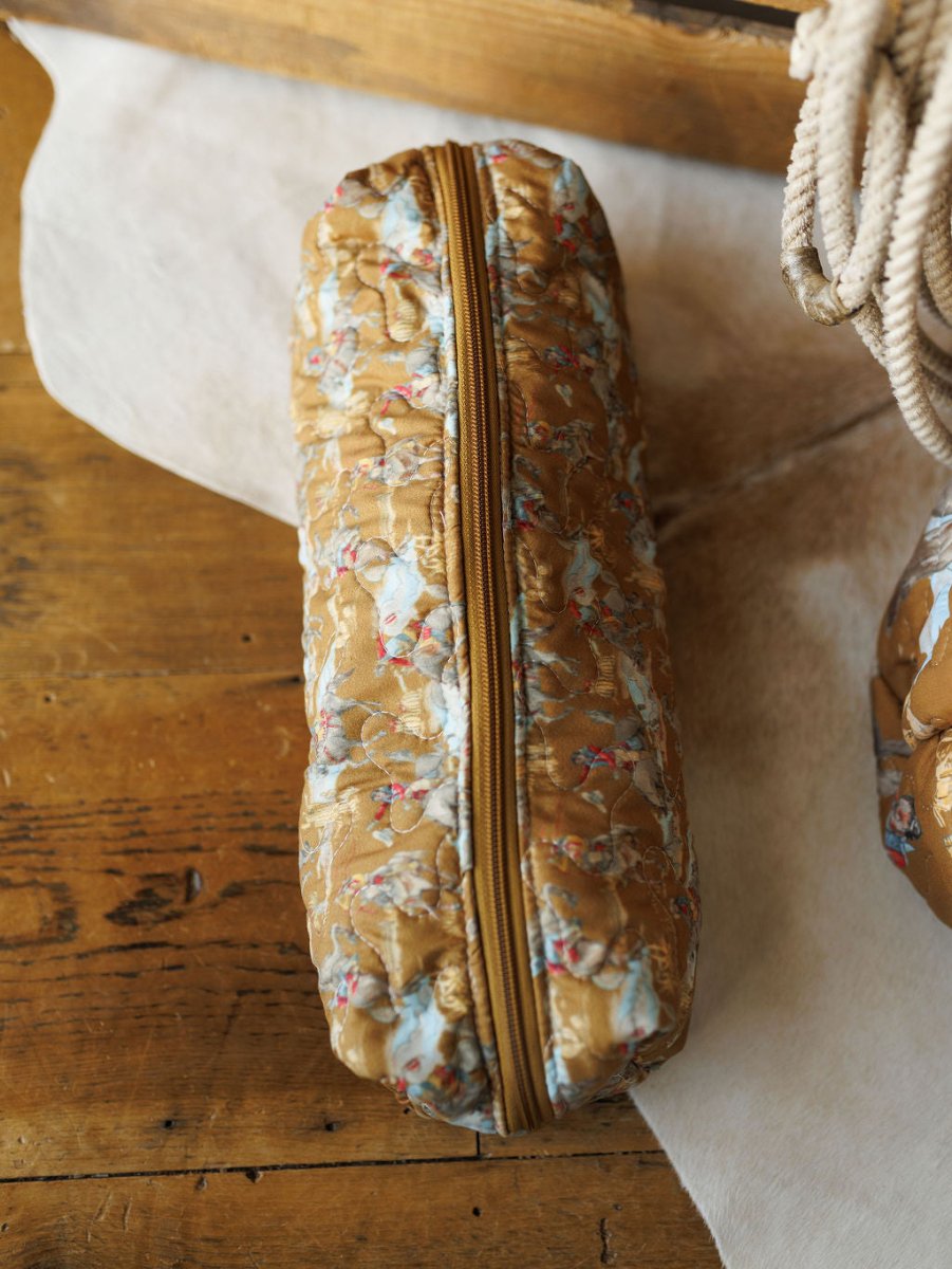 A cylindrical quilted pouch from Savannah Sevens western life&style's The Big Sky Collection, with a gold zipper and brown fabric featuring light blue floral and cherub designs, rests on a wooden surface.