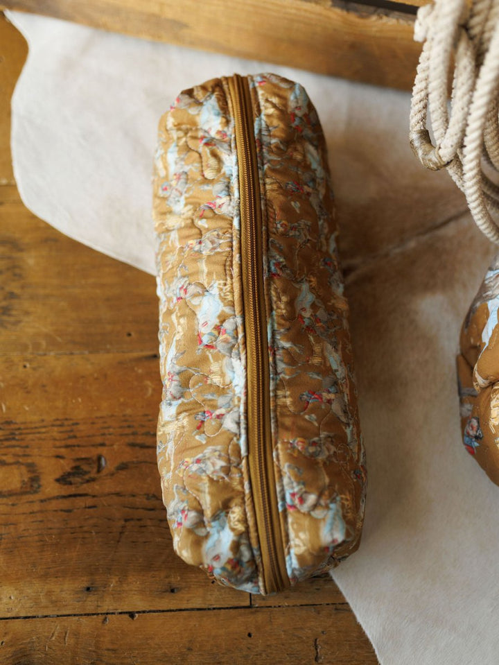 A cylindrical quilted pouch from Savannah Sevens western life&style's The Big Sky Collection, with a gold zipper and brown fabric featuring light blue floral and cherub designs, rests on a wooden surface.