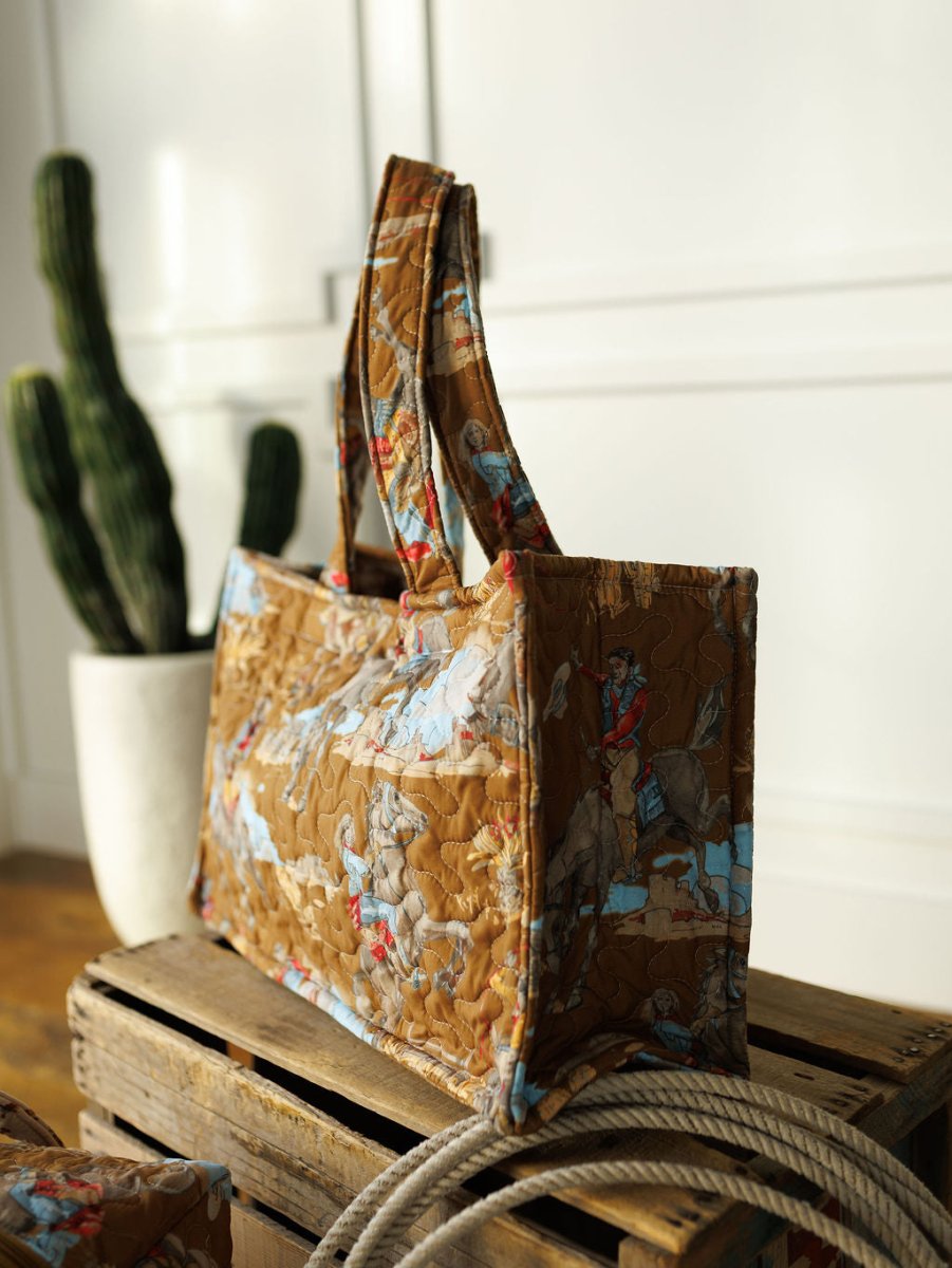 The Big Sky Collection quilted tote bag by Savannah Sevens western life&style, with brown patterns of horses and people, sits on a wooden crate beside a coiled rope and a tall cactus against a white wall.