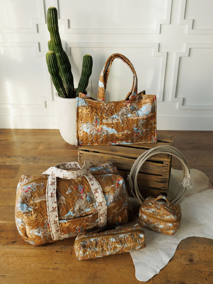 Four brown patterned quilted bags of varying sizes from The Big Sky Collection by Savannah Sevens western life{&}style are shown on a wooden floor with a white rug, crate, and potted cactus. Each bag features handles or straps.