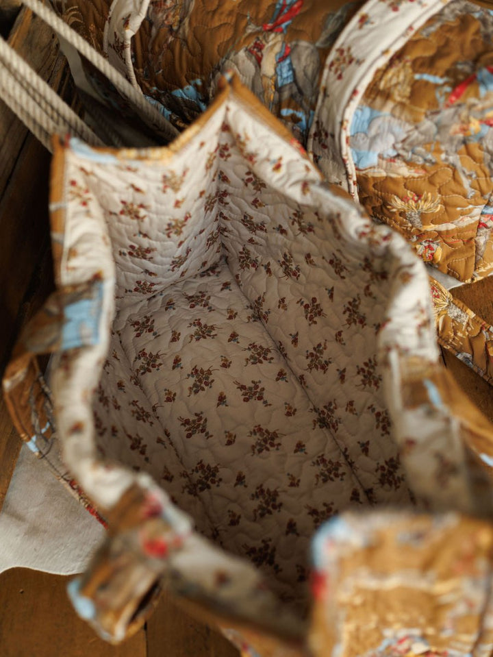 An open quilted bag from The Big Sky Collection by Savannah Sevens western life&style, shown from above with a floral lining. The exterior features quilted patches in warm tones with blue and red accents, resting on a wooden surface.