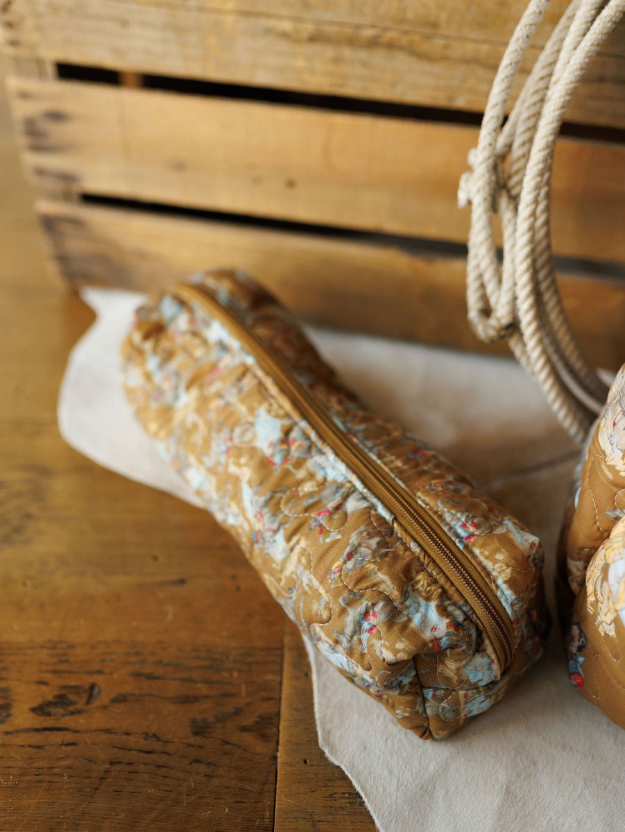 A quilted, floral-patterned pencil case from the Big Sky Collection by Savannah Sevens western life&style rests on a wooden surface near a light cloth and a rustic crate.