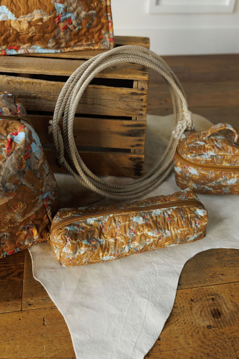 The Big Sky Collection by Savannah Sevens western life&style features textured brown and blue patterned travel bags displayed on a white hide rug next to a coiled rope and wooden crate atop a rustic wooden floor.