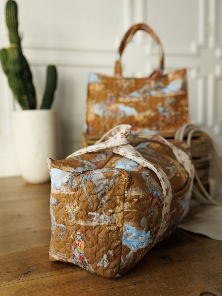 A quilted bag set from The Big Sky Collection by Savannah Sevens western life&style sits on a wooden table, displaying vibrant patterns. A matching handbag stands upright nearby next to a tall green cactus in a white pot.