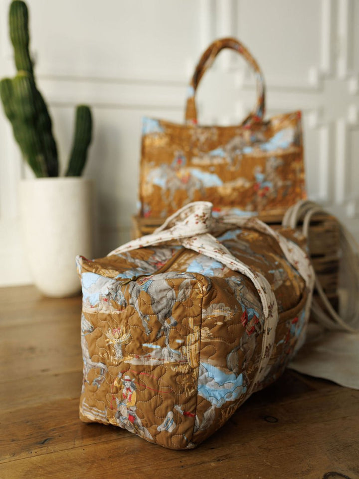 A quilted bag set from The Big Sky Collection by Savannah Sevens western life&style sits on a wooden table, displaying vibrant patterns. A matching handbag stands upright nearby next to a tall green cactus in a white pot.
