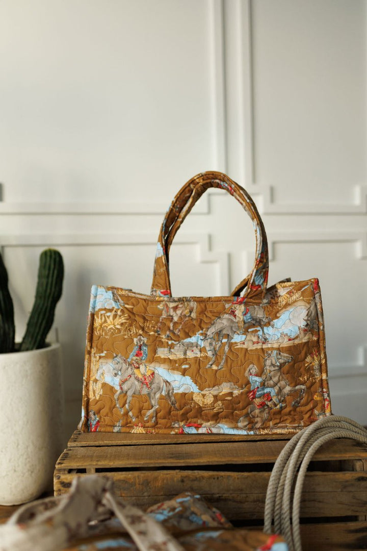 A brown patterned tote from The Big Sky Collection by Savannah Sevens western life&style, featuring cowboy and horse illustrations, rests on a wooden crate near a potted cactus and rope against a white paneled wall.