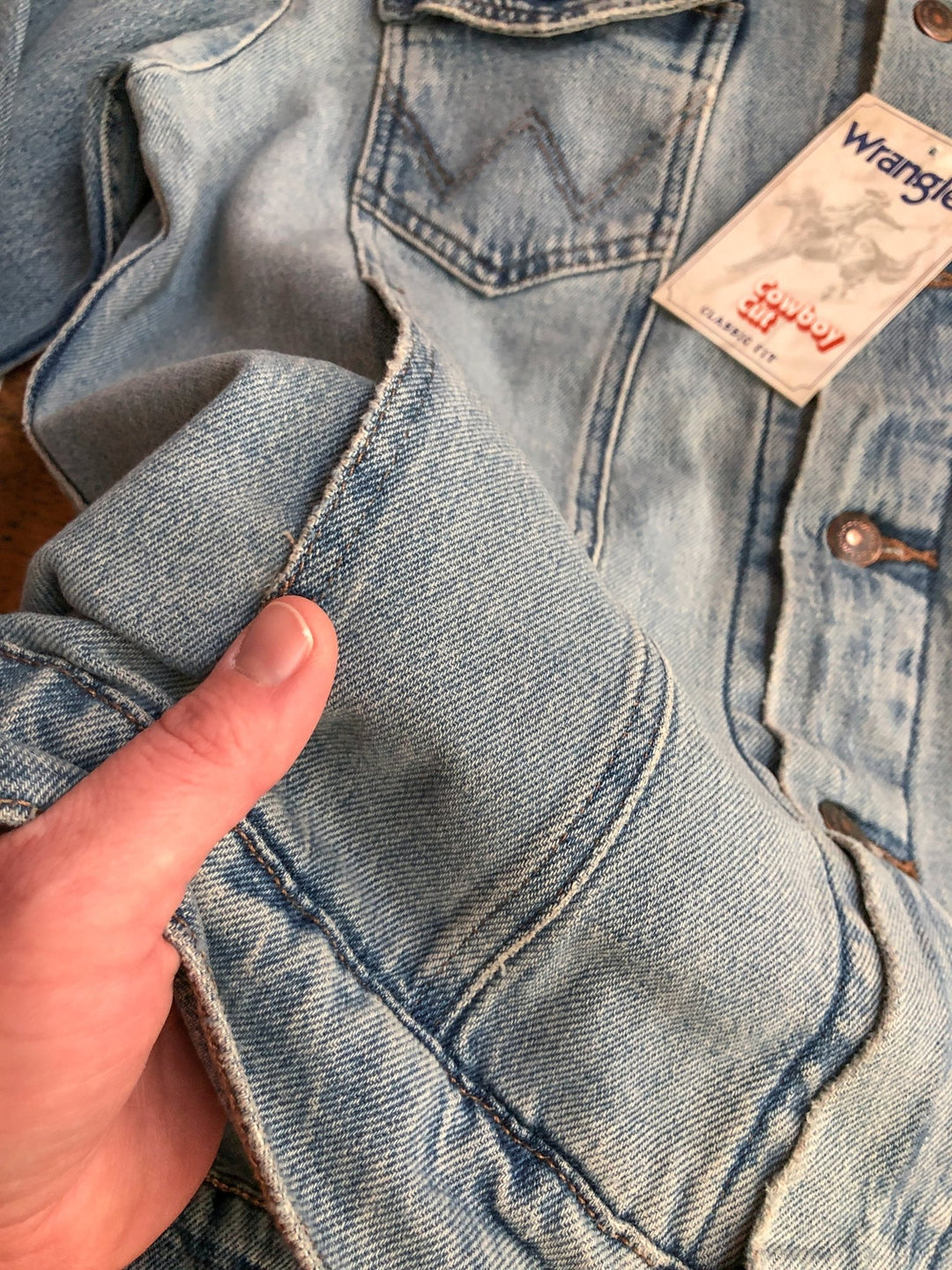 A person holds the cuff of The Boden men's light blue denim jacket by Savannah Sevens western life&style, highlighting the stitching, texture, a chest pocket, and classic button-up style.