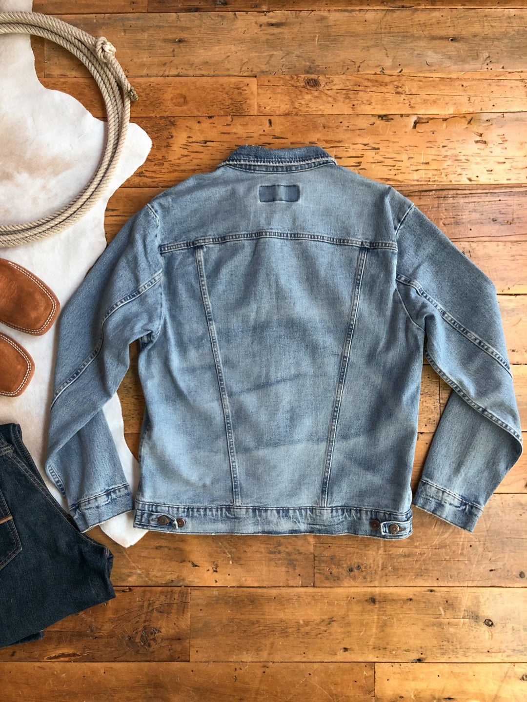 A light blue Boden {Men’s} jacket by Savannah Sevens western life{&}style is laid back side up on a wooden floor with denim jeans, brown boots, a coiled rope, and part of a white cowhide rug nearby.