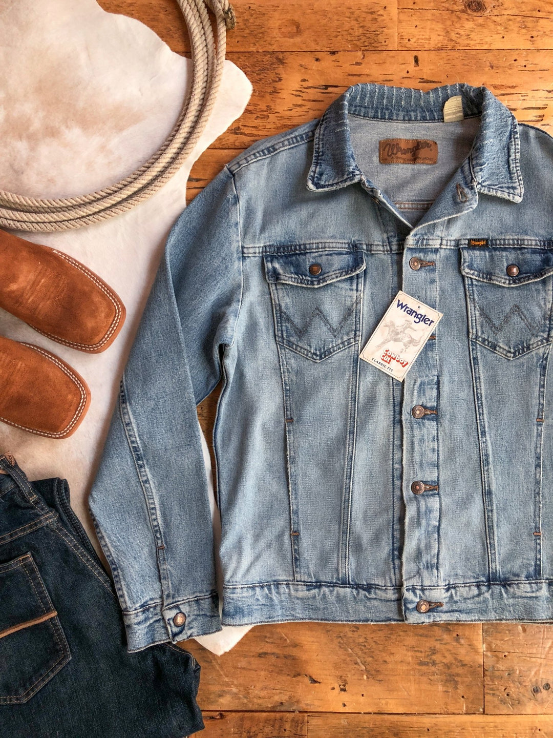 A light blue Savannah Sevens western life&style The Boden {Men's} jacket with tags is laid on a wooden floor beside brown leather boots, dark jeans, and a white animal hide rug.