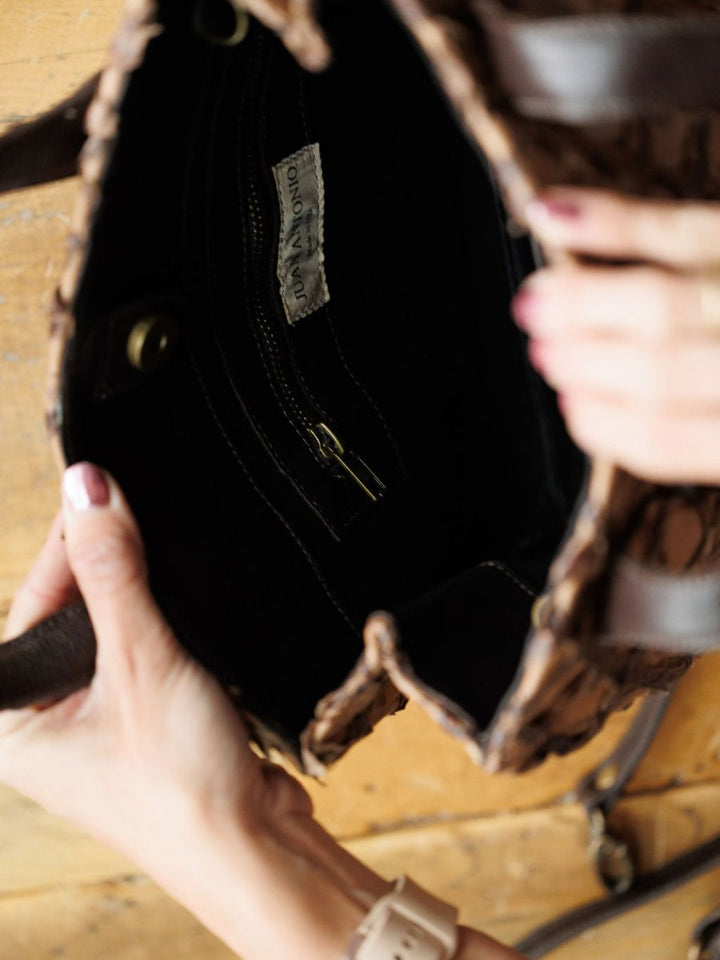 A person holds open The Brazil handbag by Savannah Sevens western life{&}style, showing its black fabric interior with a zippered pocket, gold zipper, and logo label. Shot from above on a wooden surface.