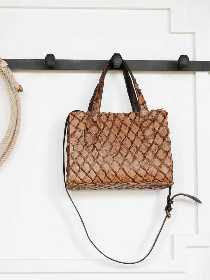 The Brazil handbag by Savannah Sevens western life{&}style, crafted from brown exotic leather with short handles and a long strap, hangs on a black wall hook beside a coiled rope against a white paneled background.
