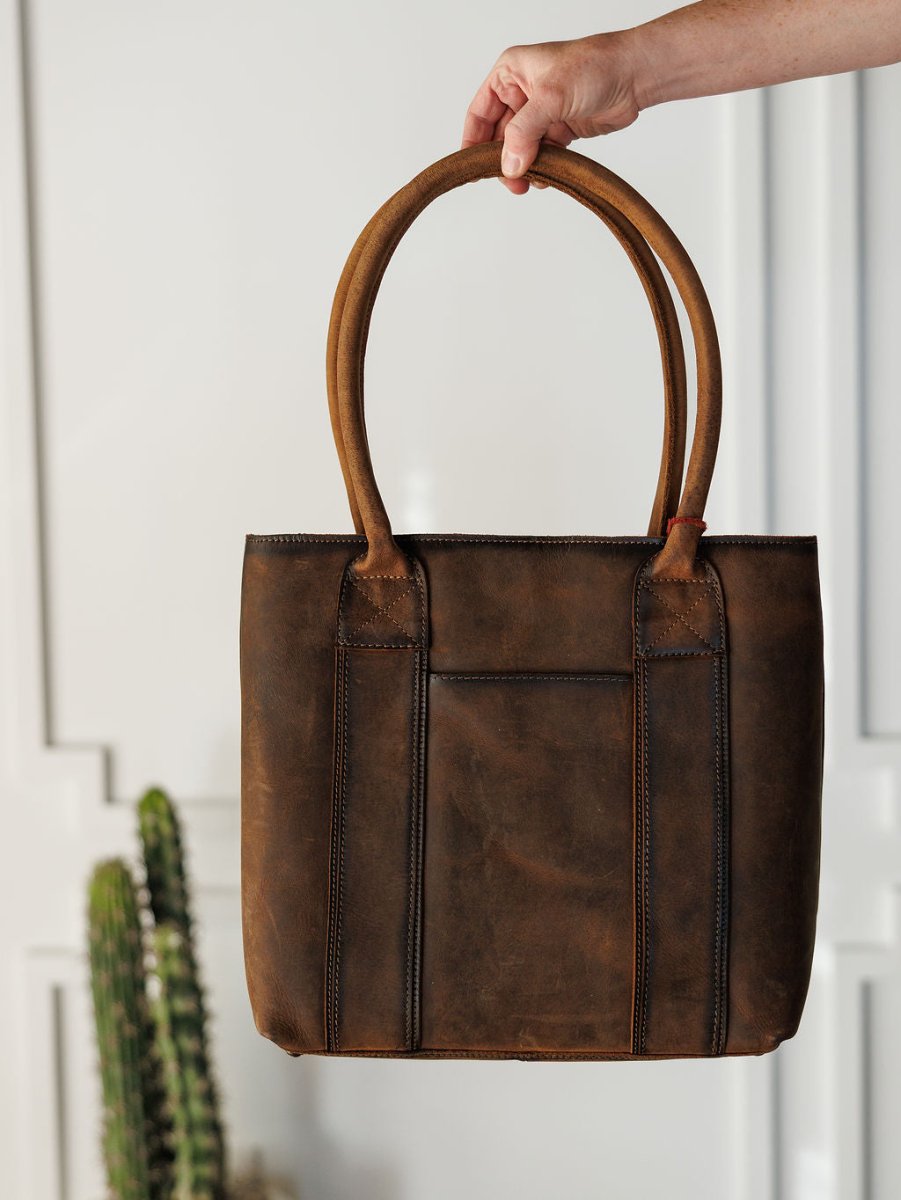 A hand holds The Broken Arrow genuine leather handbag with double handles by Savannah Sevens western life{&}style in front of a white wall and a tall cactus, emphasizing its classic western aesthetic.