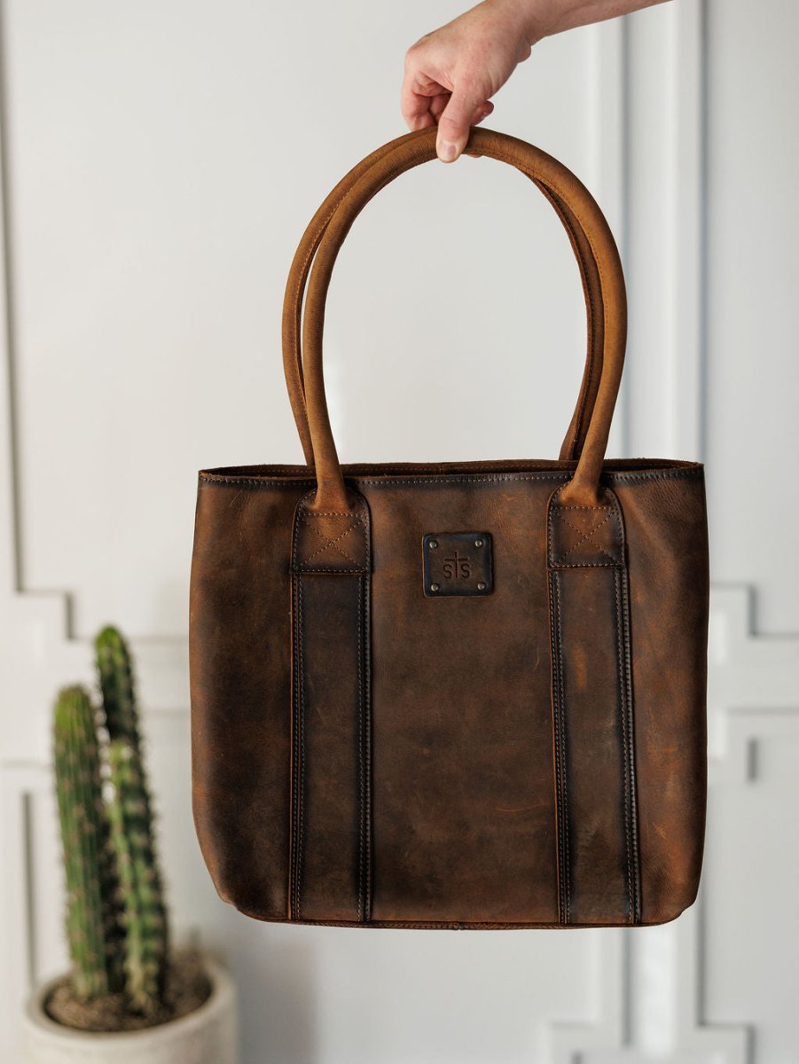 A hand holds up The Broken Arrow handbag by Savannah Sevens western life{&}style, featuring double handles and a stitched square logo. In the background, a potted cactus rests against a white wall with decorative molding.