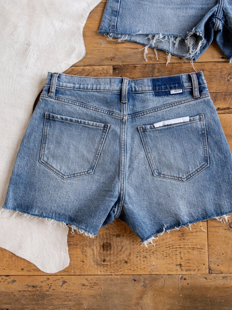 The Brynn High Rise Denim Shorts by Savannah Sevens western life{&}style, featuring a medium wash and raw cut frayed hem, are laid flat on a wooden surface next to a white cowhide rug and another pair of denim shorts.