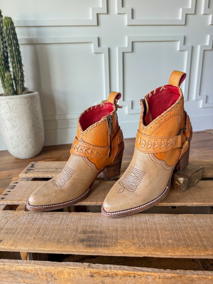 The Cadillacs by Savannah Sevens western life{&}style—tan and brown leather ankle boots with decorative stitching and a red interior—rest on a wooden pallet, with a tall cactus and white-paneled wall accenting their classic western style.